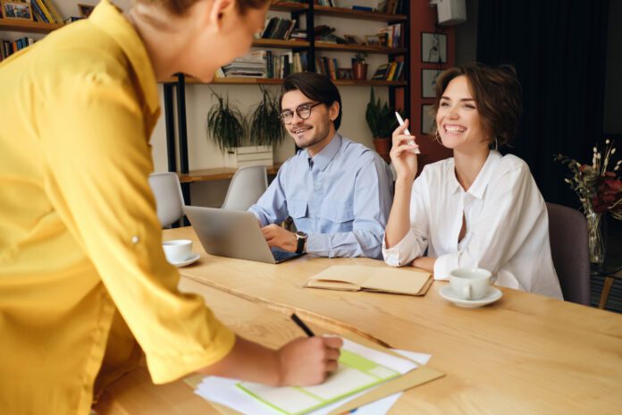 Group of cheerful business colleagues sitting at the desk with laptop and papers while happily talking at work in modern office